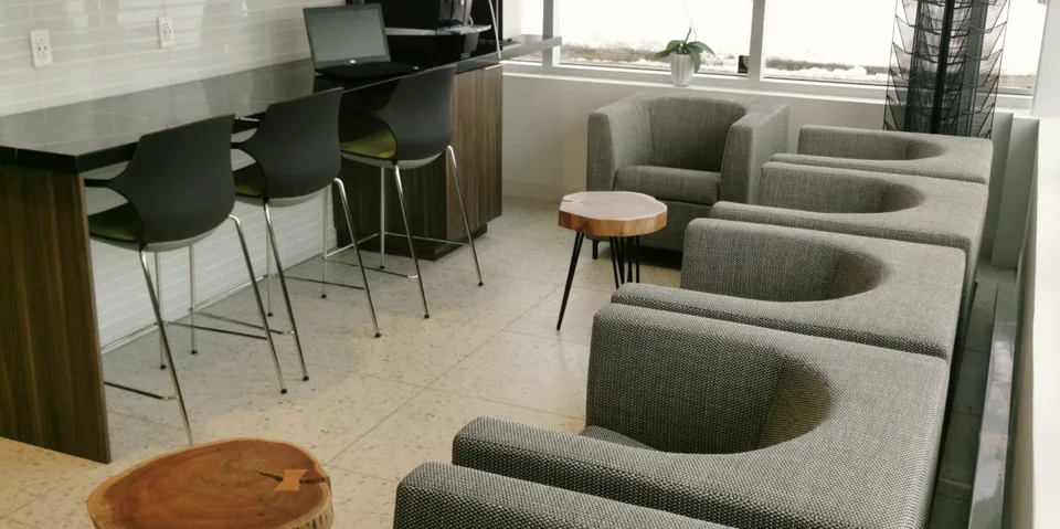 Salle d’attente avec fauteuils gris et tables en bois près des fenêtres.
